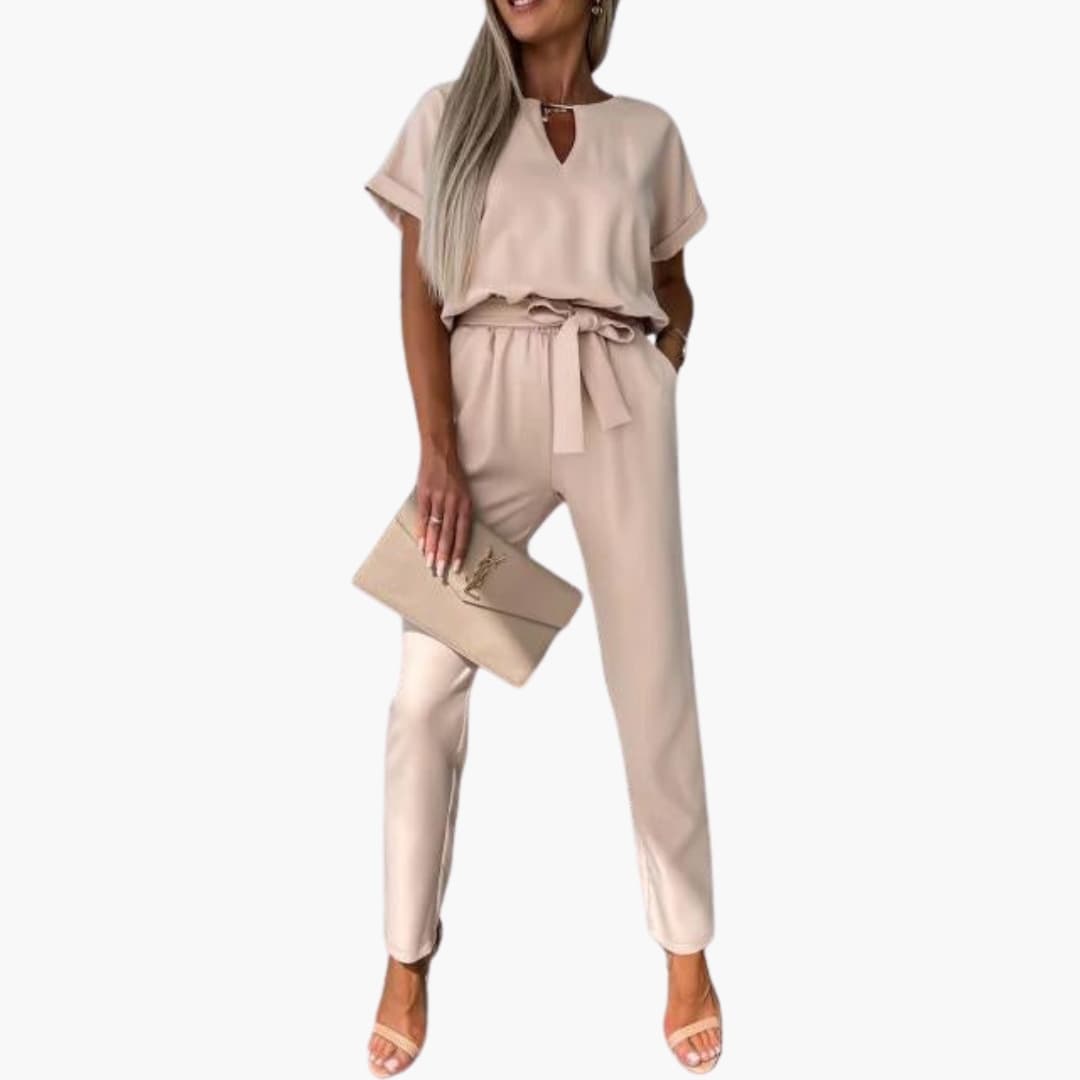 Women's V-neck jumpsuit with waist belt in beige, by Wilfred Smith, paired with a beige logo clutch